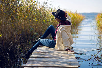 Woman in a black hat and sweater sits on the old pier to the bushes by the lake
