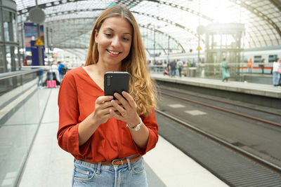 Smiling attractive brazilian traveler passenger buys ticket online with smartphone in train station.