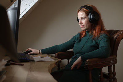 Middle age woman listening to music in big headphones near computer at home. creating play list