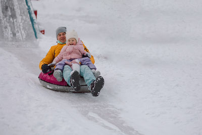 Little girl in pink with her dad roll down ice slide outdoors on winter snowy day. joy and fun 