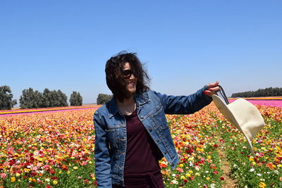 Young woman holding hat on flower field against clear sky