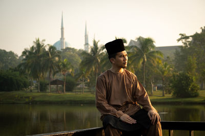 Man wearing traditional clothing while sitting on railing against lake during sunset