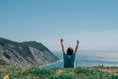 An adult woman sits on a mountain barefoot, raising her hands to the sky and the sun on a sunny day