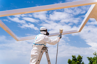 Low angle of anonymous employee in protective uniform and mask standing on ladder and using air brush to paint frame structure against cloudy sky on sunny day