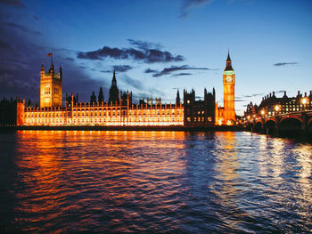 Illuminated buildings by river against sky in city at dusk