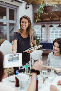 Smiling female owner giving menu cards to customer sitting in restaurant