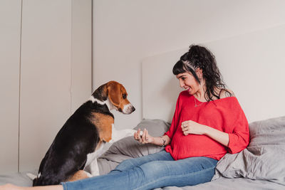 Pregnant young woman shaking hands with dog while sitting on bed at home