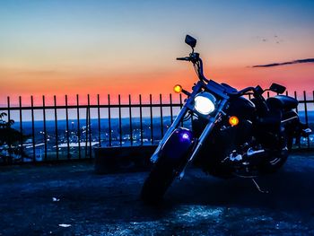 Motorcycle parked on field by railing against sky during sunset