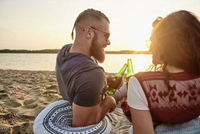 Smiling couple with beer bottles sitting at beach against sky during sunset
