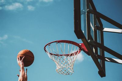 Cropped hands aiming basketball into hoop against sky