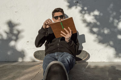 Young man reading book while sitting with skateboard on footpath