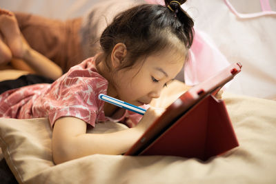 Asian adorable kid girl is lying on the floor in living room at home and writing on tablet 