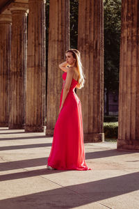 Full length side view of young woman in pink evening gown standing at colonnade