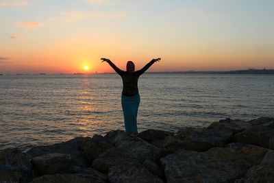Rear view of woman standing on rock at sea shore against sky during sunset