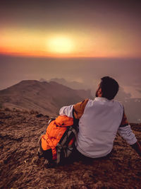 Rear view of man sitting on land against sky during sunset