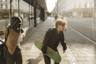 Teenage boy with skateboard laughing while standing by female friend on footpath in city