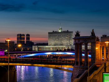 Illuminated bridge over river against buildings at dusk