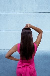 Back view of unrecognizable slim brunette with gorgeous long hair dressed in elegant pink outfit standing with hand on head against blue wall