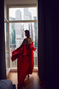 Woman in a red bathrobe stands at a large window in an apartment opposite a skyscraper