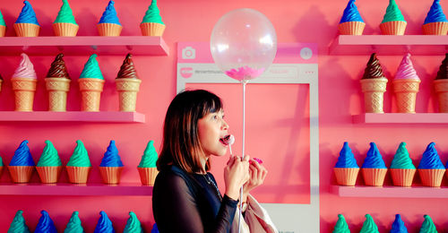 Woman holding balloon and eating cake pop while standing against wall