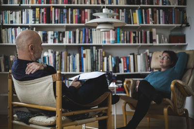 Male therapist discussing with female mature patient against bookshelf at home office