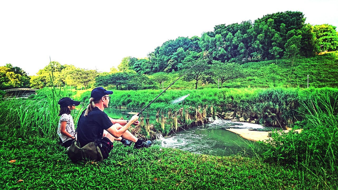 lifestyles, leisure activity, tree, men, sitting, green color, grass, person, clear sky, relaxation, growth, water, rear view, plant, nature, full length, casual clothing, tranquility