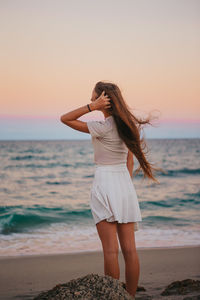 Rear view of woman standing at beach against sky during sunset