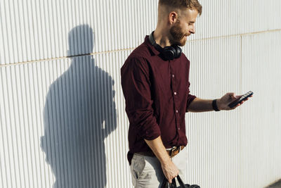 Businessman with headphones using mobile phone while standing by white wall