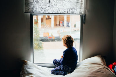 Full length of boy looking through window while sitting on bed at home