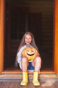 A cheerful girl with long hair is sitting on a doorstep, holding a pumpkin with eyes and mouth 