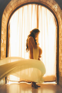 Side view of naked pregnant woman standing by window at home