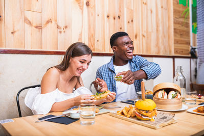 Positive young multiracial friends eating tasty dishes and burgers while gathering in modern restaurant