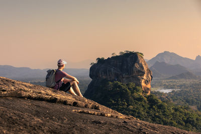 Man sitting on rock against sky during sunset