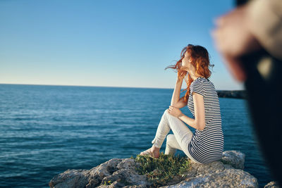 Side view of young woman sitting on rock by sea against sky