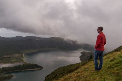 Side view of man standing on mountain by lake against cloudy sky