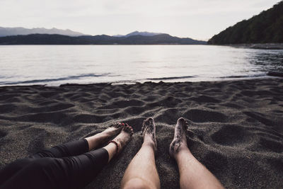 Low section of couple sitting at beach against sky