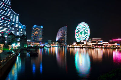 Illuminated ferris wheel by river and buildings against sky at night