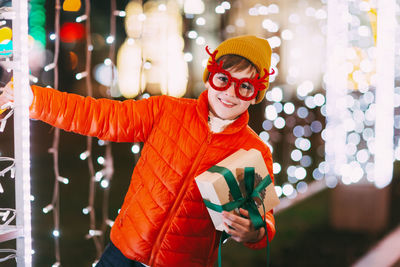 A cheerful boy in an orange jacket and funny glasses walks through the evening city 