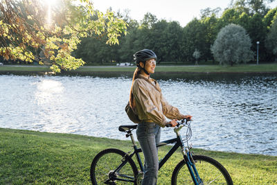 Smiling woman with bicycle standing by lake in park