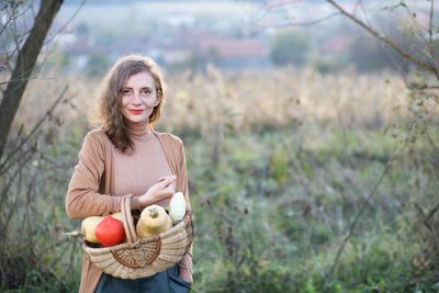 Portrait of smiling woman holding fruits in basket standing against field