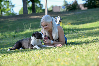 Young blonde woman is lying on lawn while her dog is licking her finger after feeding with goodies.