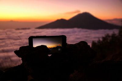 Silhouette of camera on rock against sky during sunset
