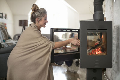 Smiling woman putting wood in fireplace at home