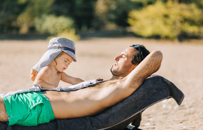 Shirtless father with daughter relaxing on lounge chair at beach