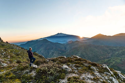 Man standing on top of the cliff in mountains at sunset enjoying sunset over erupting volcano etna.