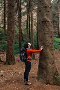 Side view of woman in backpack touching tree trunk and looking up while exploring forest on summer day