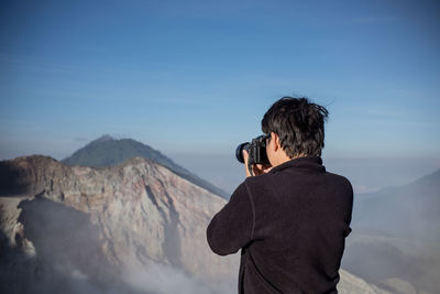 Rear view of man photographing mountains against blue sky