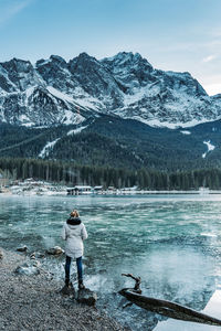 Rear view of woman standing at lakeshore against snowcapped mountains
