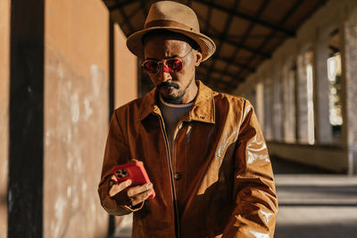 Serious bearded african american male in hat and stylish sunglasses surfing modern cellphone while standing on sunny street in city