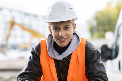 Smiling teenage construction trainee wearing hardhat and reflective clothing at site